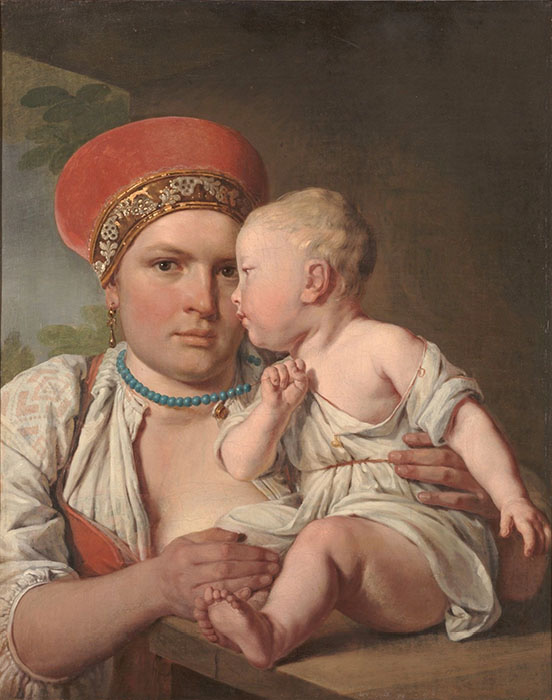 Woman with bare breast holds sitting child on the table in from of her; she is wearing a dangling earrings, a blue stone bead necklace, and a cylindrical hat with embroidered trim 