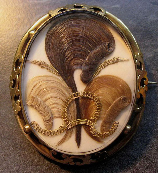 artfully arranged locks of hair: one blond, one brunette, one auburn, under glass in a metal setting on a pin