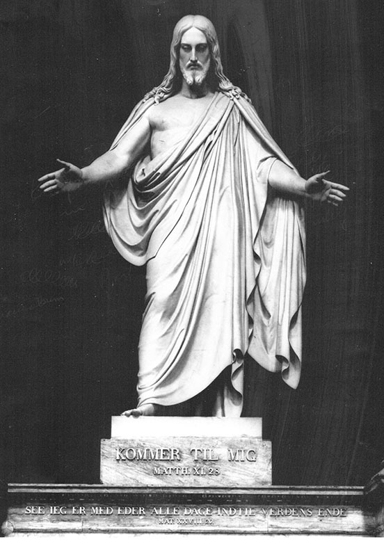 statue of christ with arms outstretched downwards, with "Kommer til mig" (Come to me) in Danish on the pedestal 