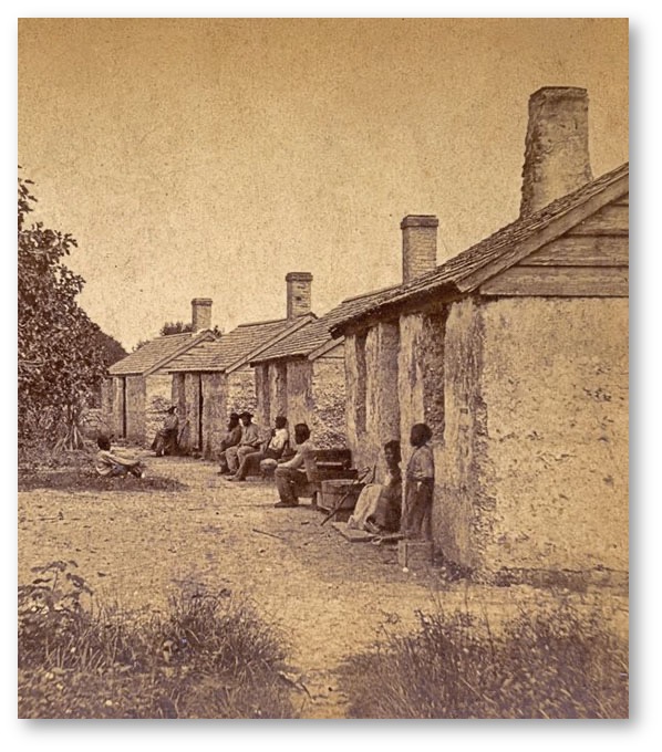 A sepia photograph of a row of shanty houses on the right, with a Black child, woman, and five men sitting spaces along the walls; on the left, tropical vegetation with one Black youth sitting in the grass